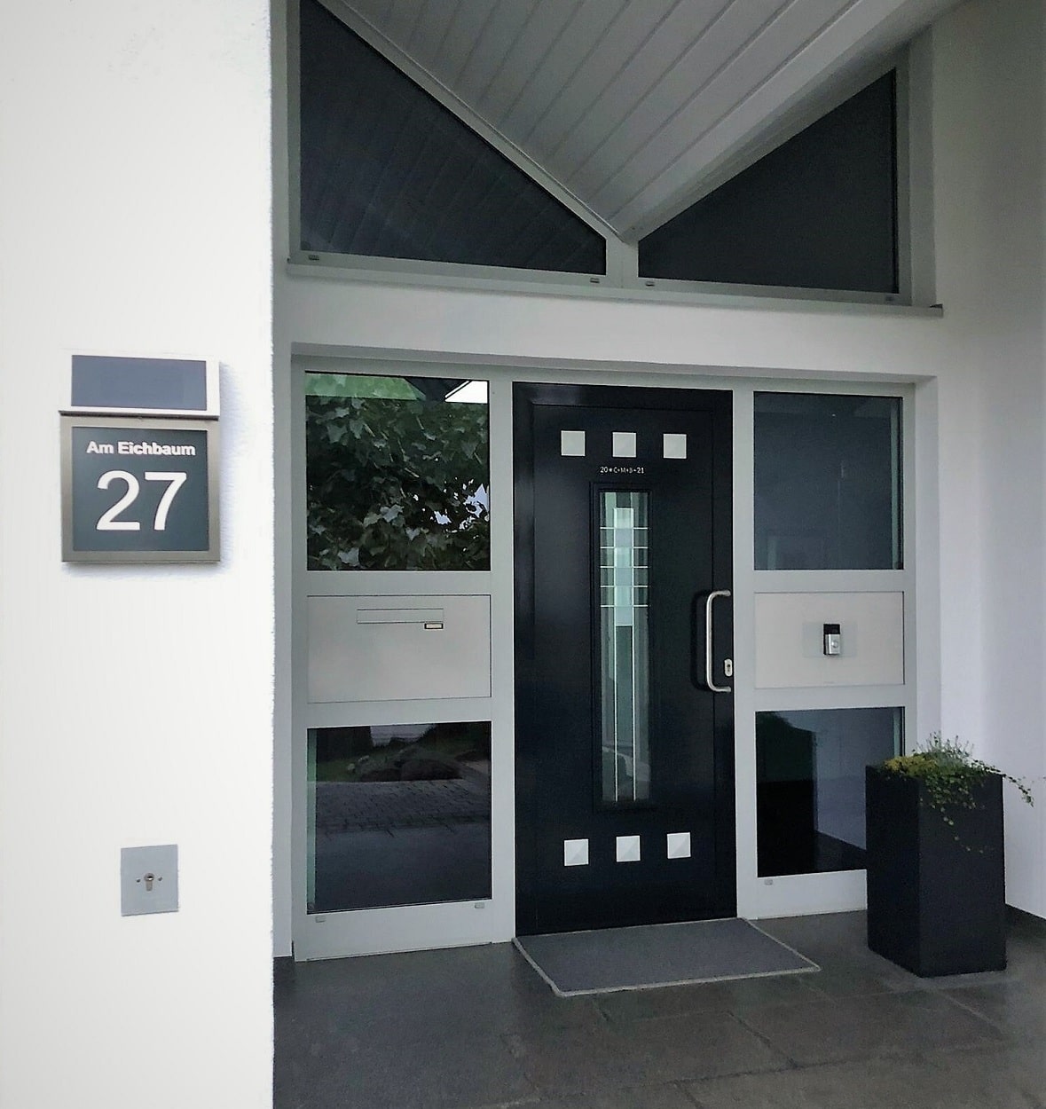 Edelstahl Solarhausnummer mit grauem Hintergrund und weißer Beschriftung in einem weiß gehaltenen Eingangsbereich neben einer dunkelgrauen Tür