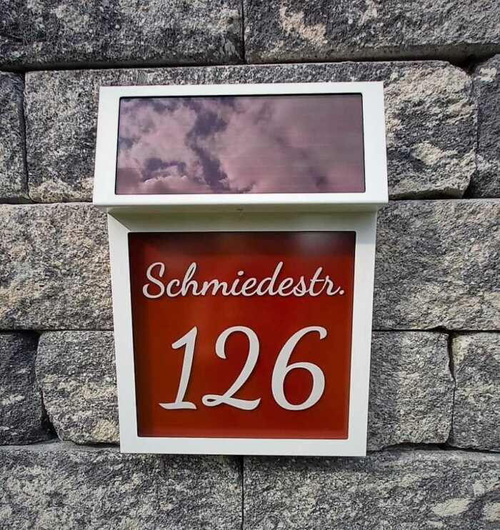 Solarhausnummer mit weißem Gehäuse und Beschriftungsfeld mit rotem Hintergrund. Verspielte Schrift mit Aufschrift Schmiedestr. 126.